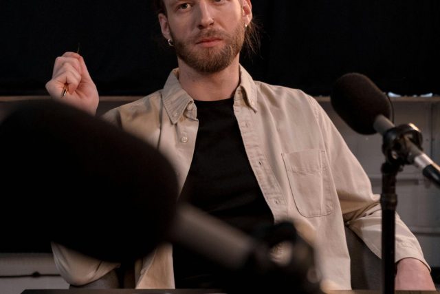 Bearded caucasian man in beige shirt podcasting in a studio with microphones.
